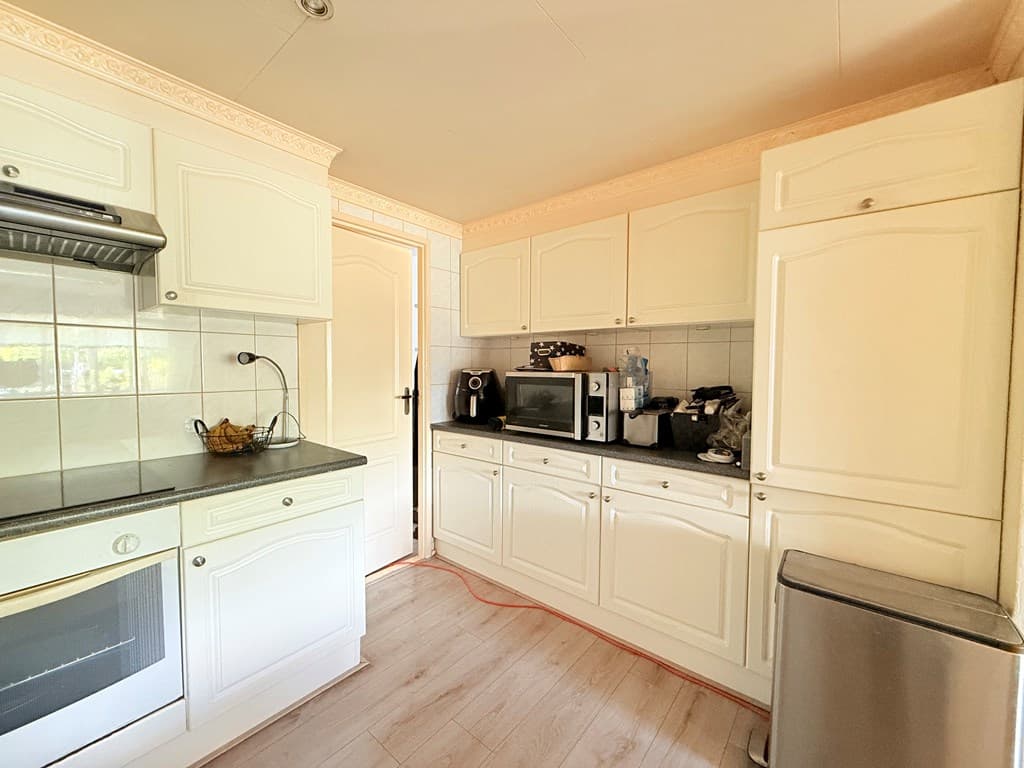 Witte keuken met boven- en onderkasten, oven, magnetron en koelkast zichtbaar, deur naar een andere ruimte aan de rechterzijd