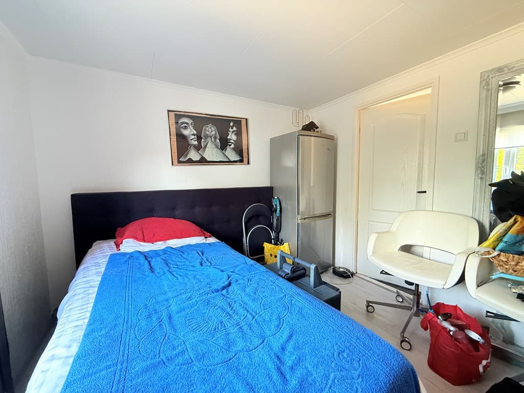 Slaapkamer met een bed, een schilderij aan de muur, een koelkast en een witte stoel naast een nachtkastje.