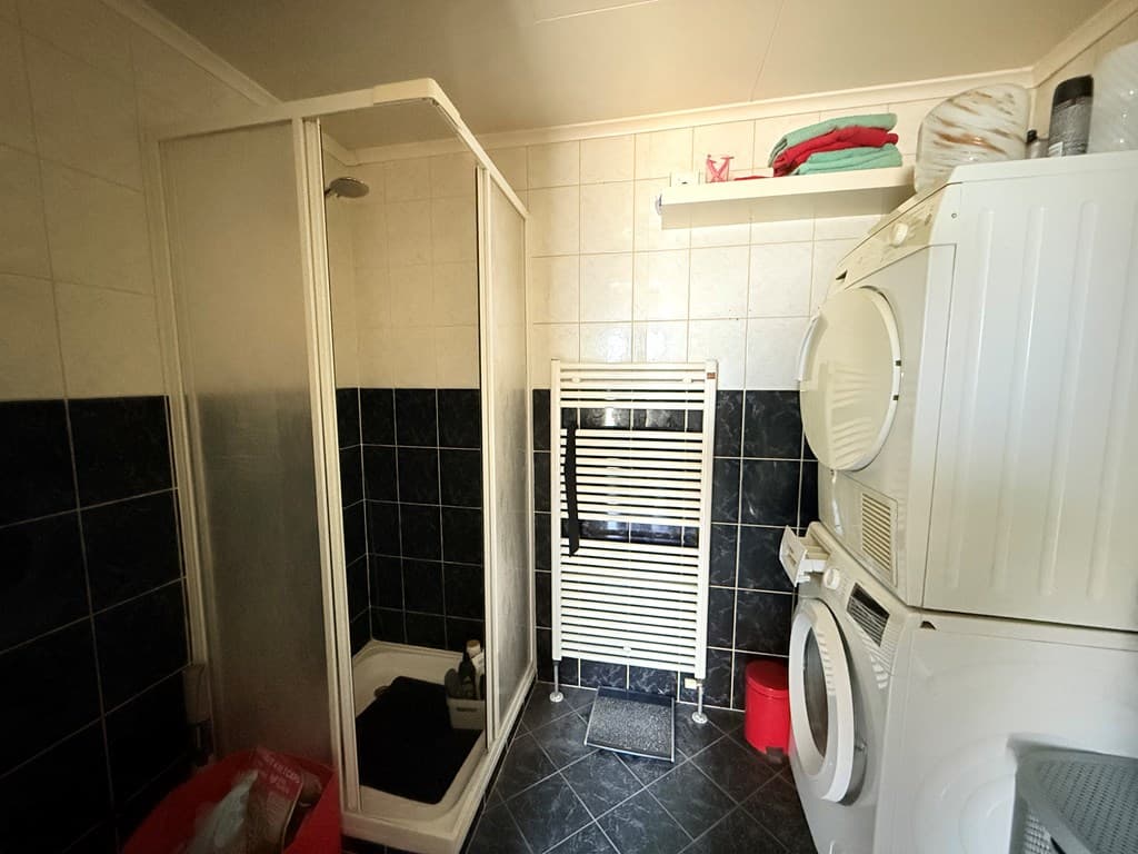 Badkamer met douchecabine, wasmachine en droger, en een handdoekradiator aan de muur.