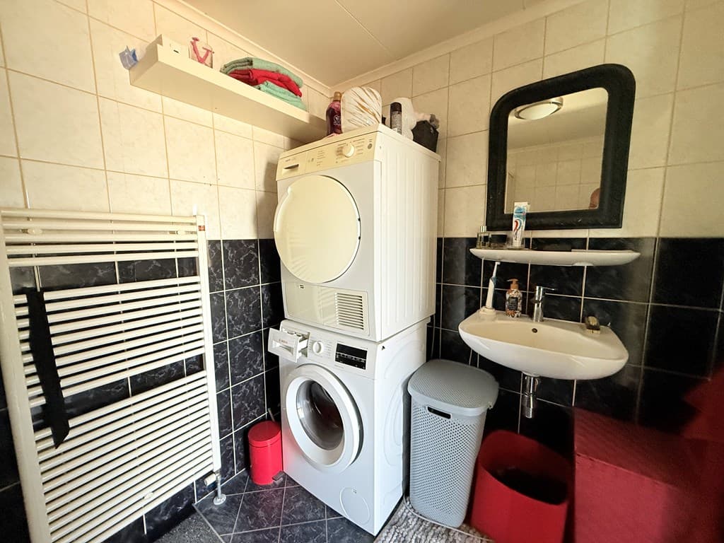 Waskamer met wasmachine, droger, wastafel, spiegel en een radiator in een overzichtelijke ruimte.