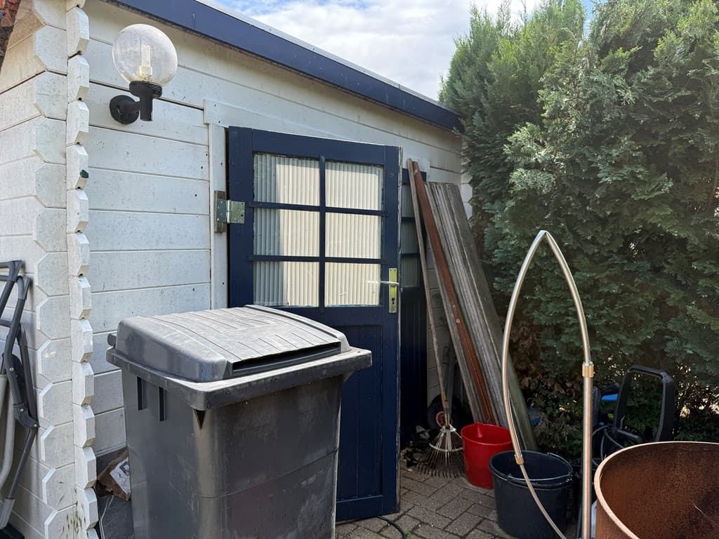 Buitenaanzicht van een schuur met een blauwe deur, vuilnisbak, tuinmateriaal en groenblijvende struiken eromheen