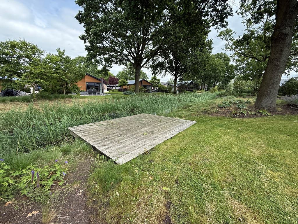 Houten aanlegsteiger bij een groene border met riet en zicht op vakantiewoningen in de achtergrond.