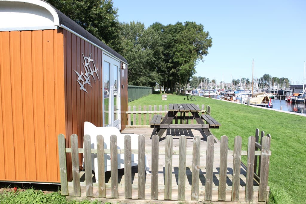 Terras met houten tafel en stoelen, omheind door een houten hek, uitzicht op de jachthaven met boten en groen.
