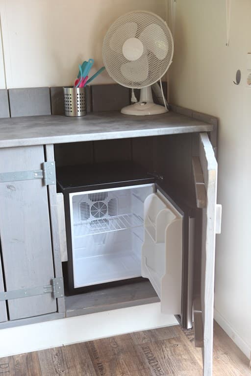 Keuken met een open kast en een kleine koelkast, daarnaast een ventilator en enkele keukengerei op het aanrecht.