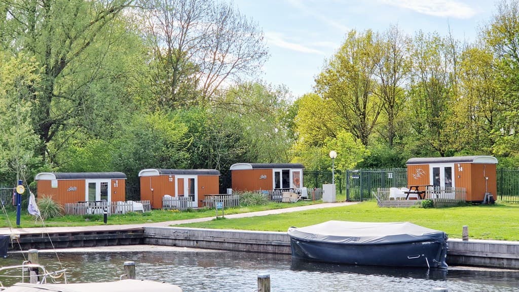 Drie vakantiehuisjes in een groene omgeving aan een jachthaven, met een aanlegsteiger en een boot in het water.