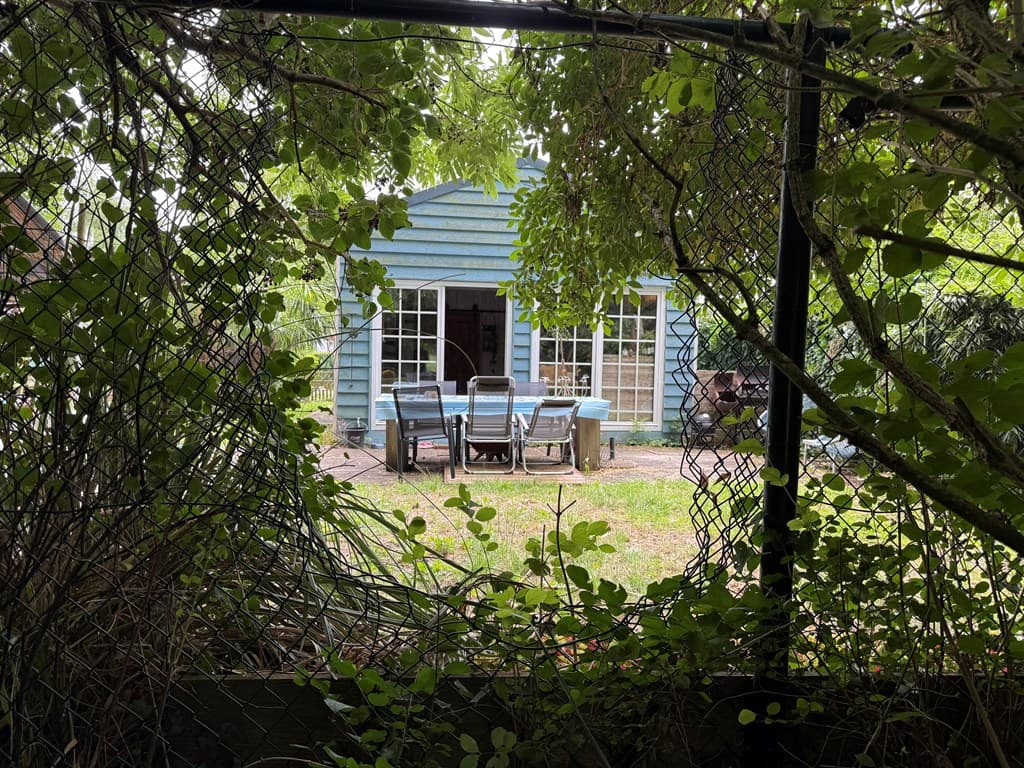 Zicht op een blauwe woning met grote ramen en een terras met stoelen, omringd door groen en een hekwerk