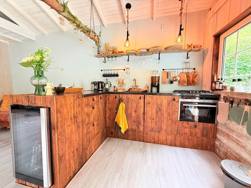 Keuken met houten kasten, moderne apparatuur en groene planten; grote ramen zorgen voor veel licht.