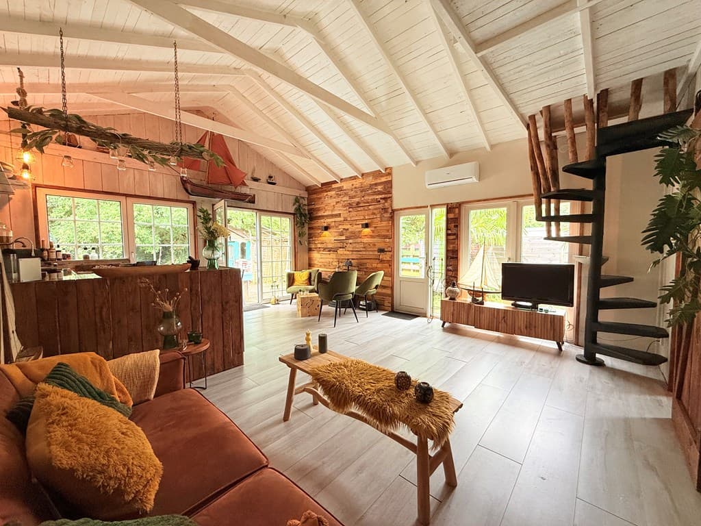 Ruime en lichte woonkamer met houten accenten, bank, eettafel en toegang tot een buitenruimte met uitzicht op de natuur.