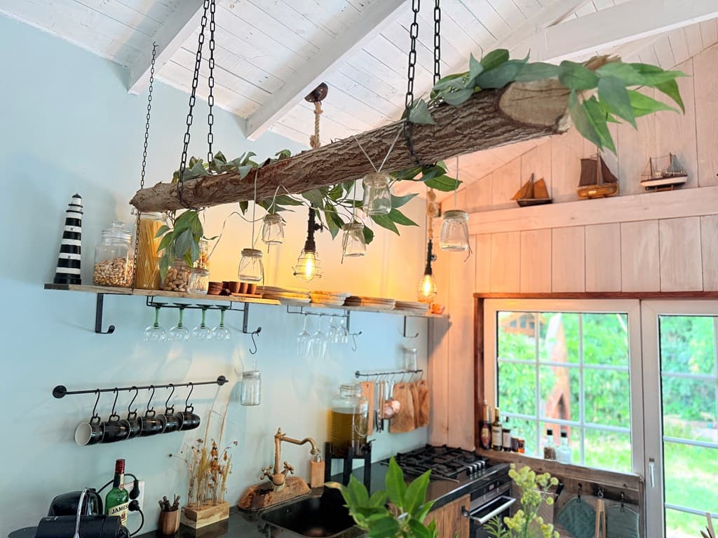Keuken met een houten balk aan het plafond, hangende glazen potten, en een open kast met maritieme decoratie.