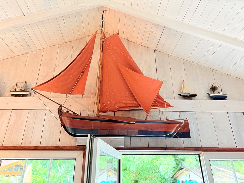 Houten zeilboot hangt aan de wand, omringd door kleinere scheepjes, met grote ramen voor natuurlijk licht.