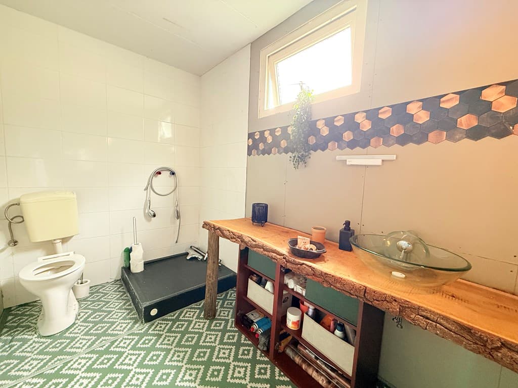 Badkamer met douche, toilet en houten wastafel met opbergruimte, betegelde muren en een venster met uitzicht naar buiten