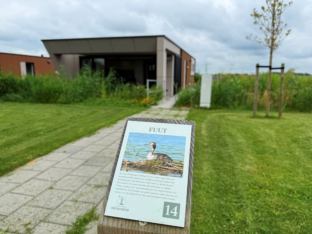 Moderne woning met groene tuin, nabij water, met informatiebord over een fuut op de voorgrond.
