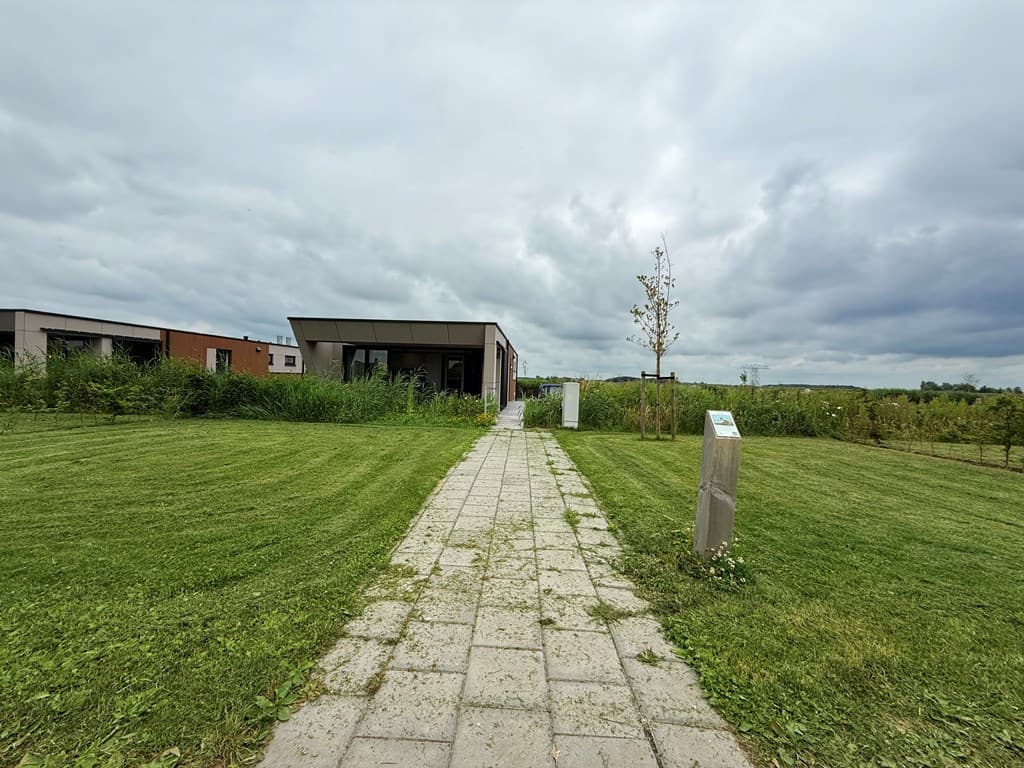 Moderne vakantiewoning met een betegeld pad, omgeven door gras en enkele bomen onder een bewolkte lucht