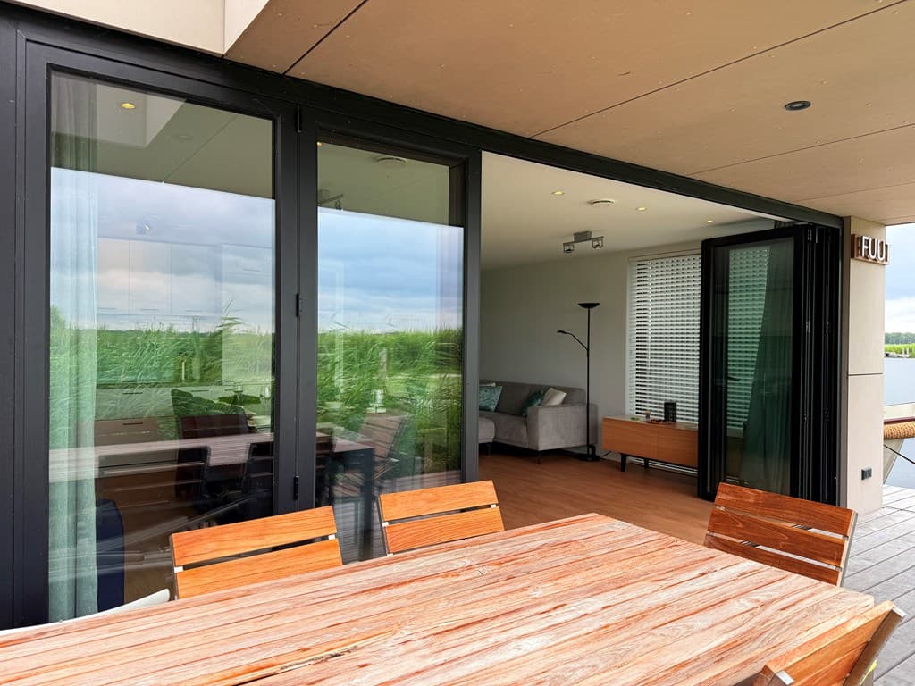 Terras met houten tafel, openslaande deuren naar woonkamer met sofa en lamp, uitzicht op groen omgeving.