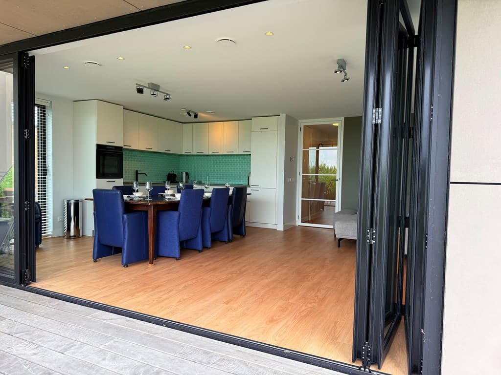 Moderne open keuken met eettafel en blauwe stoelen, zicht op het terras door openslaande deuren.