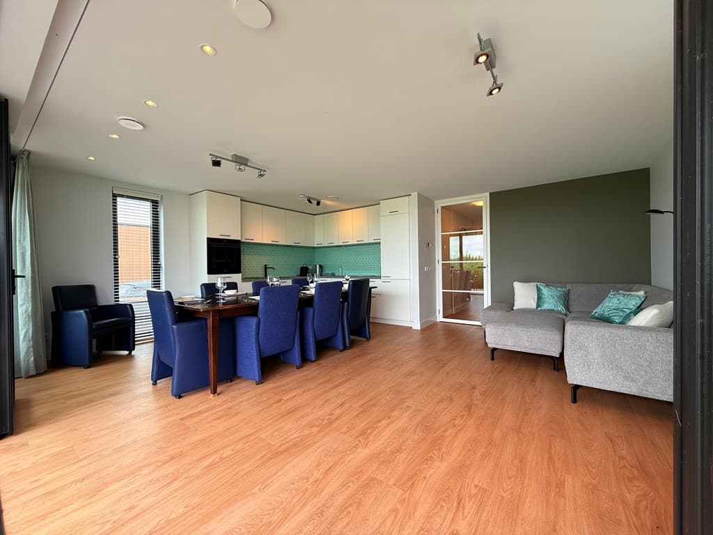 Moderne woonkamer met open keuken, grote eettafel en zithoek met bank en kussens, veel natuurlijk licht en houten vloer.