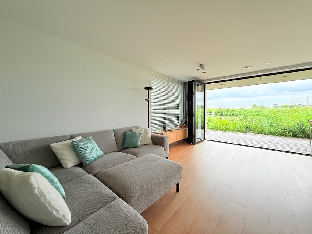 Hedendaagse woonkamer met grijs bankstel en groene kussens, met uitzicht op groene omgeving door grote glazen deur.