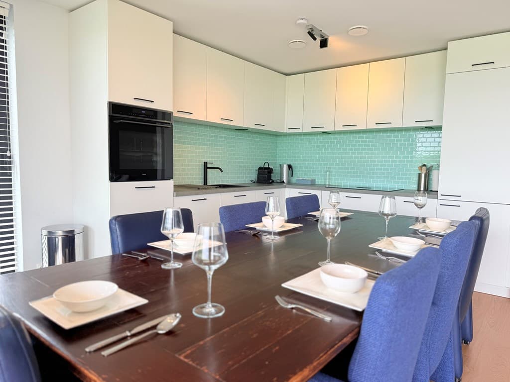 Moderne keuken met een lange houten eettafel, blauwe stoelen en glazen servies, groene wandtegels en inbouwapparatuur