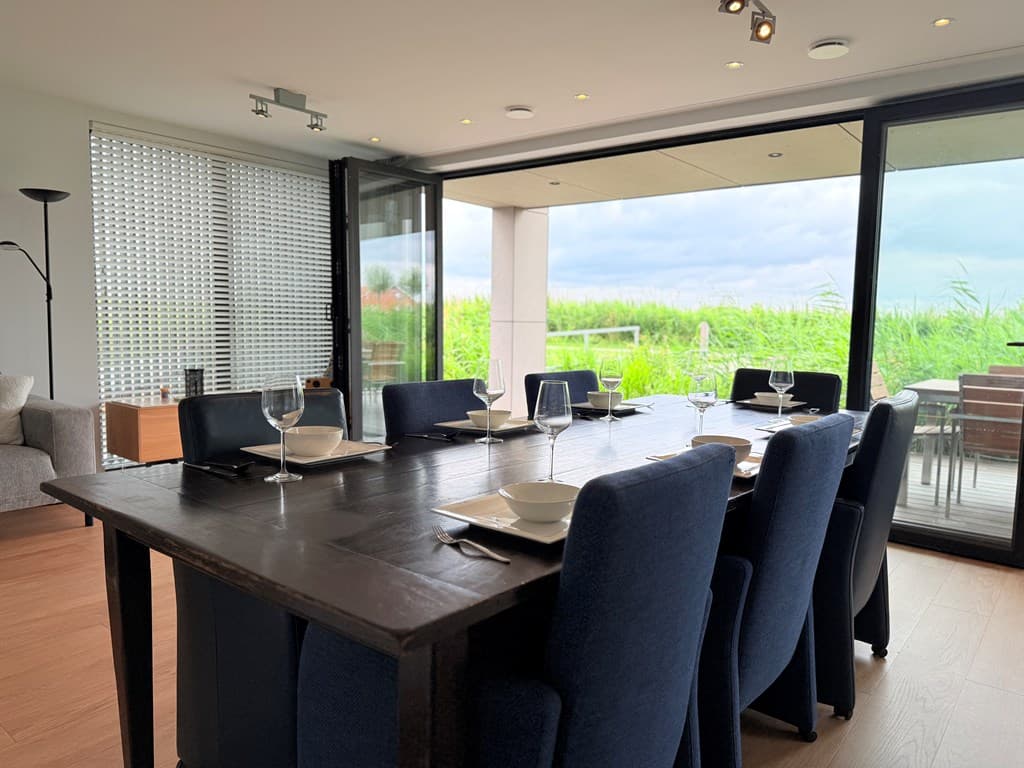 Eetkamer met grote tafel, blauwe stoelen en uitzicht op het groen door grote glazen schuifdeuren.