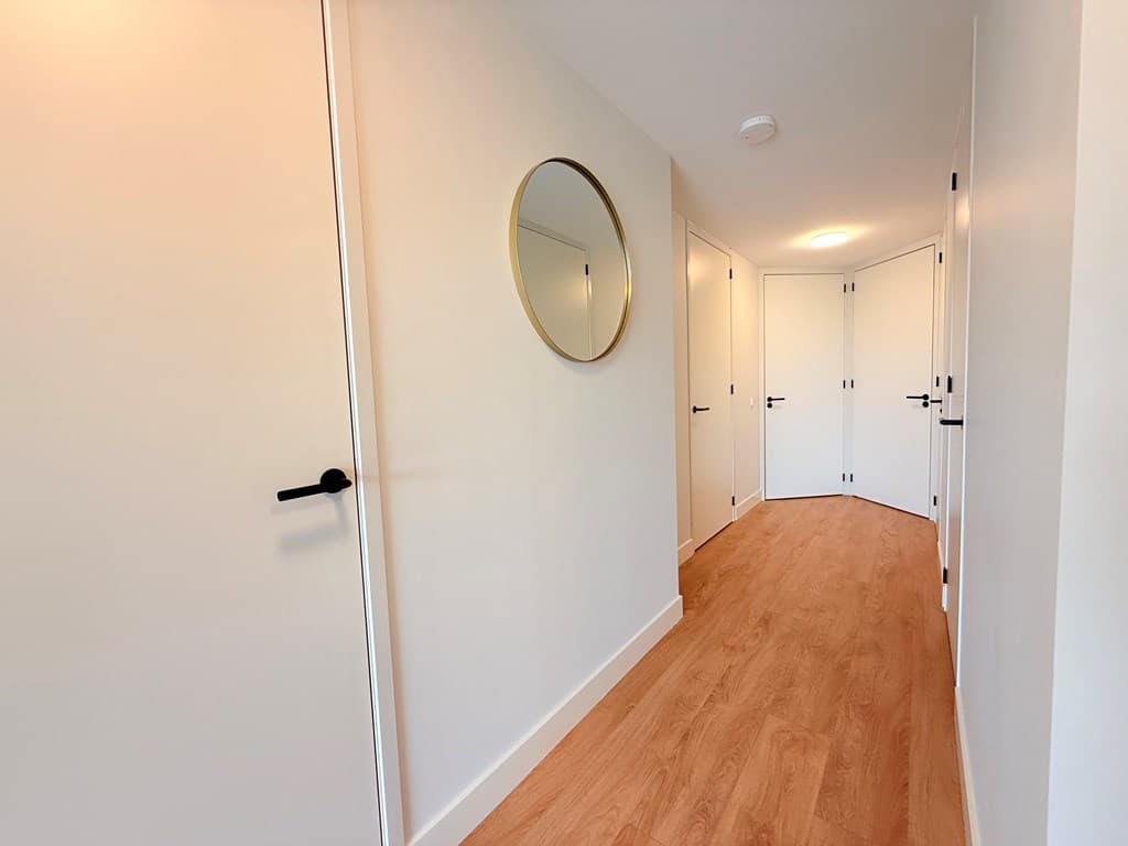 Lichte hal met houten vloer, witte wanden en een spiegel aan de muur; deuren naar verschillende ruimtes zichtbaar.