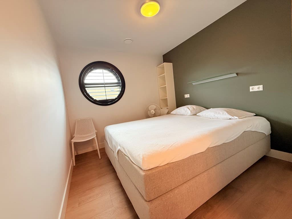 Slaapkamer met een tweepersoonsbed, ronde raampartij en een stoel tegen de muur.