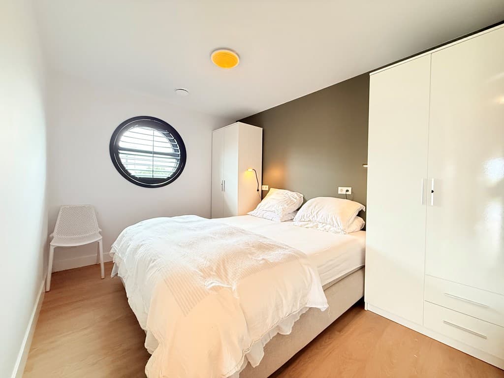 Sfeervolle slaapkamer met een rond raam, een tweepersoonsbed en een kledingkast tegen de muur.