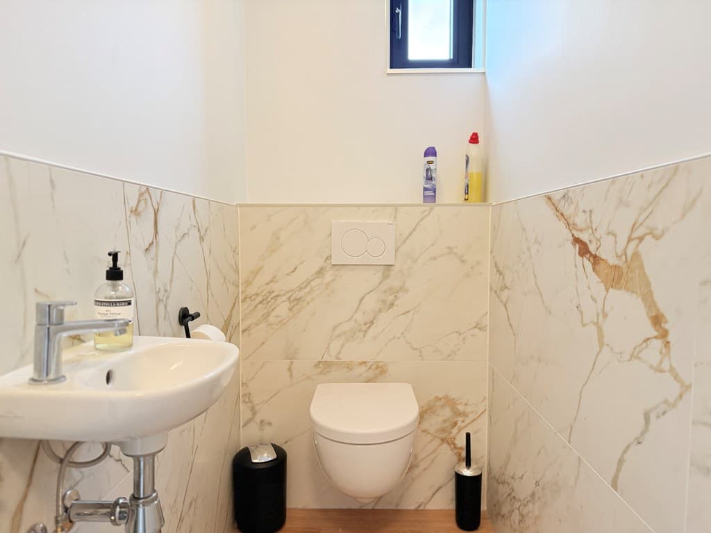 Wc met marmeren wandtegels, een toilet, wastafel en enkele schoonmaakproducten op de planken