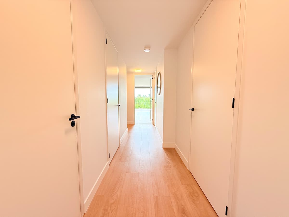 Lange gang met lichte wanden en een houten vloer, deuren aan beide zijden en natuurlijk licht aan het einde van de gang.