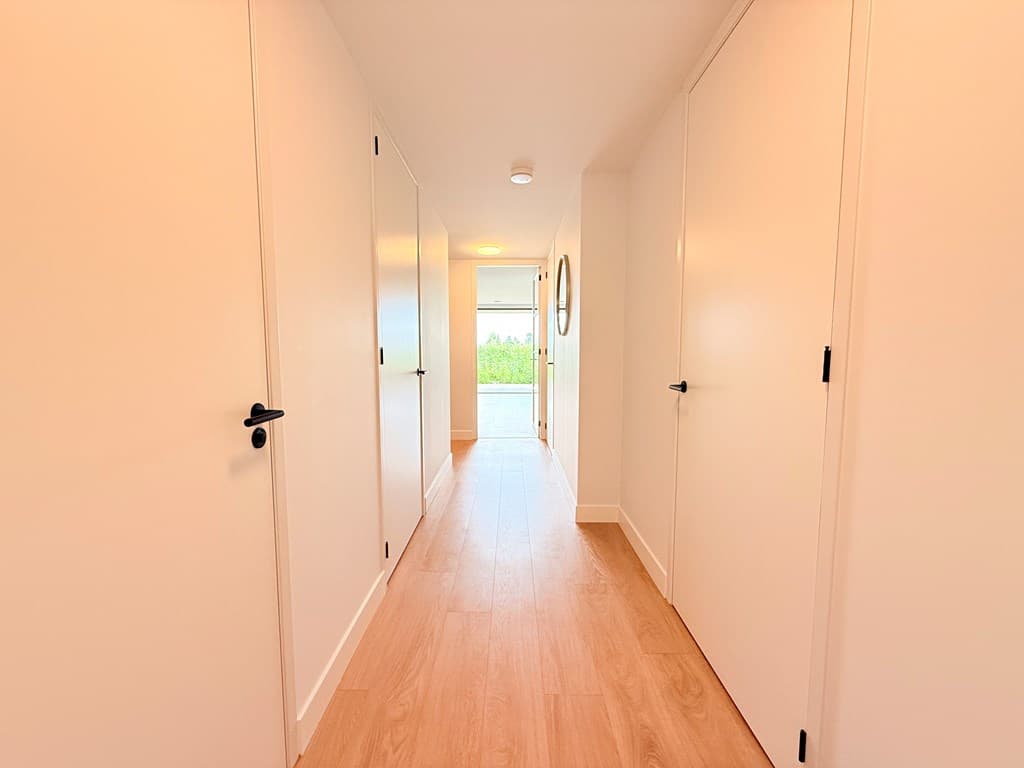 Lange gang met lichte wanden en een houten vloer, deuren aan beide zijden en natuurlijk licht aan het einde van de gang.