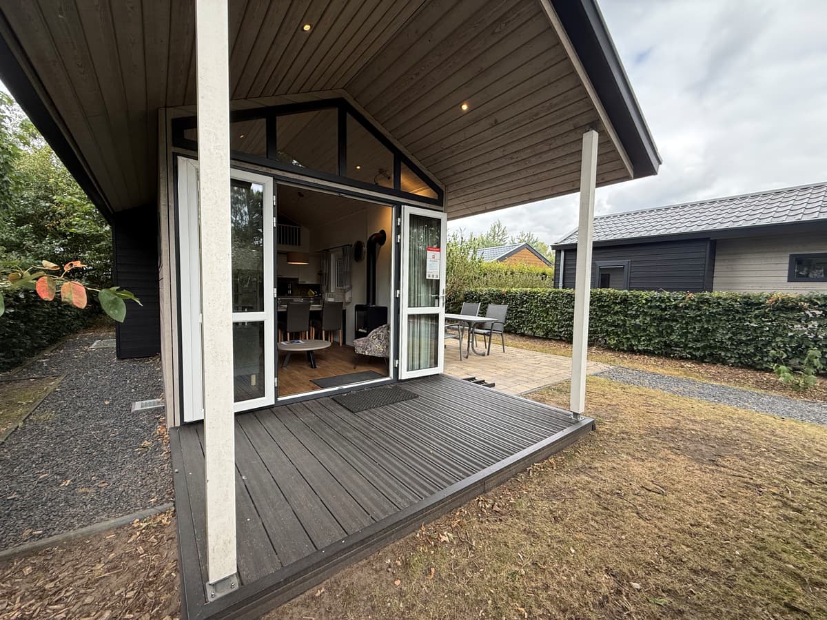 Vooraanzicht van een vakantiewoning met terras en dubbele openslaande deuren, omringd door groen en een pad naar de naastligg
