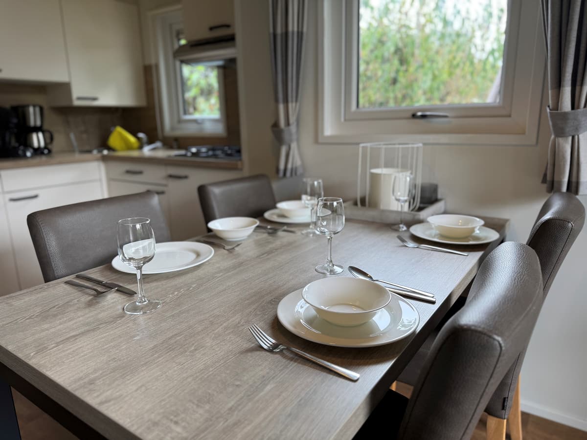 Eettafel gedekt met borden, glazen en bestek met uitzicht op een keukenruimte en ramen met uitzicht op groen.