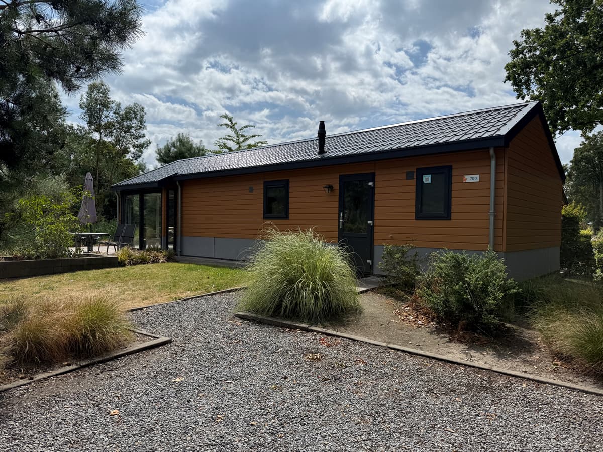 Houten vakantiewoning met een donkere dakbedekking, omgeven door groen en grindpaden, en een terras met parasol zichtbaar.
