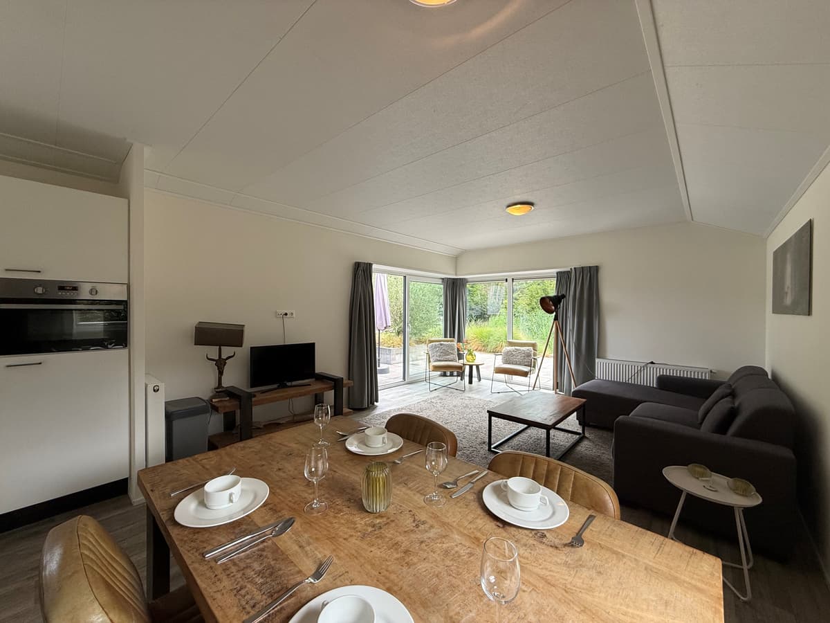 Huiselijke woonkamer met eettafel, bank en toegang tot een terras met groene omgeving en grote ramen.