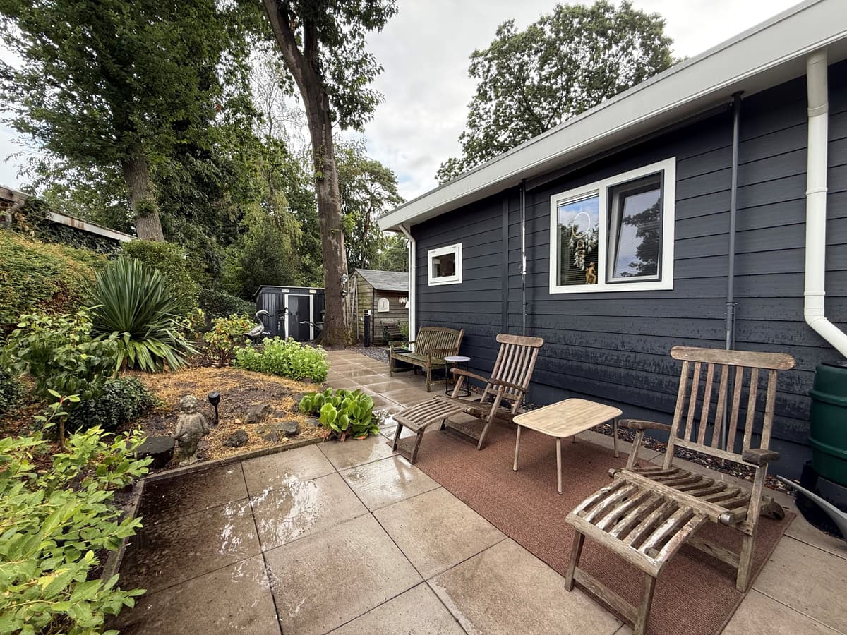 Terras met houten stoelen en tafel, omlijst door groen plantenbed en bomen, aan de achterkant van een bungalow.
