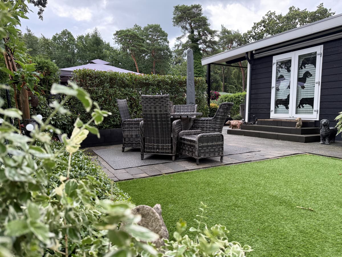 Terras met tuinmeubels van wicker en kunstgras, omringd door struiken en bomen bij een recreatiewoning in Harderwijk