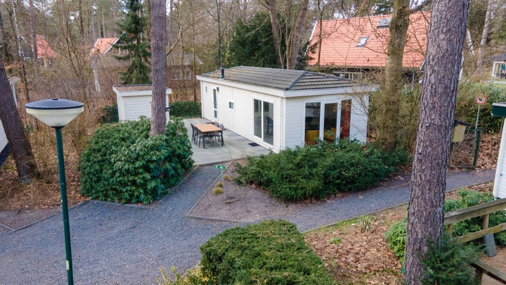 Vakantiehuisje omringd door bomen en planten, met een terrasje en een pad van grind eromheen.