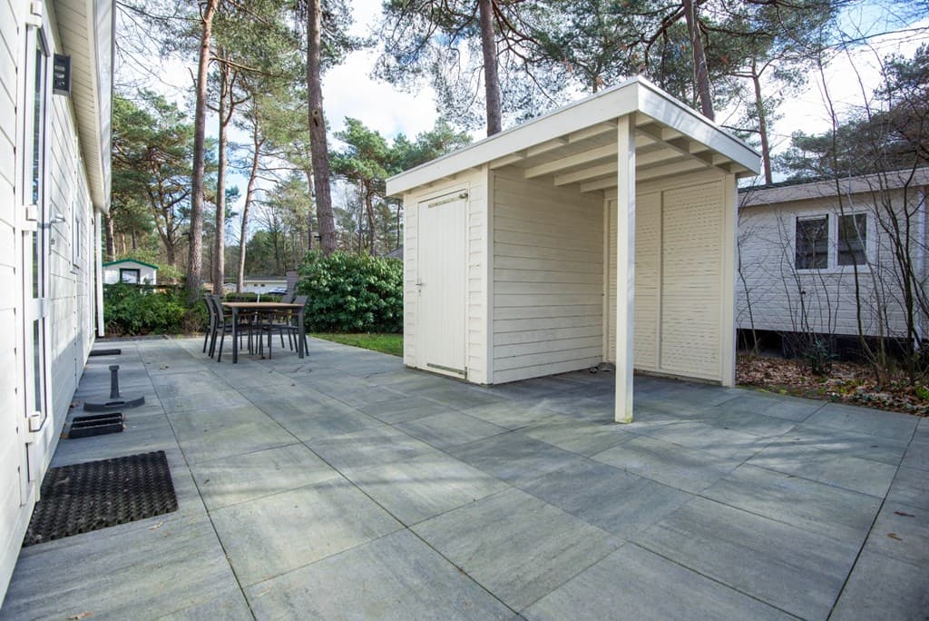 Terras met grijze tegels, een houten berging en een eettafel met stoelen, omringd door bomen en groen.