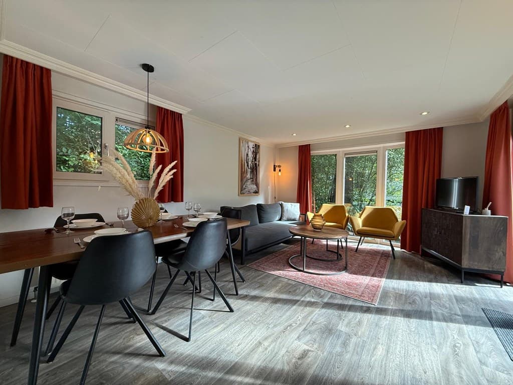 Woonkamer met eettafel, fauteuils, tv, grote ramen en decoratieve elementen zoals een vaas met droogbloemen.