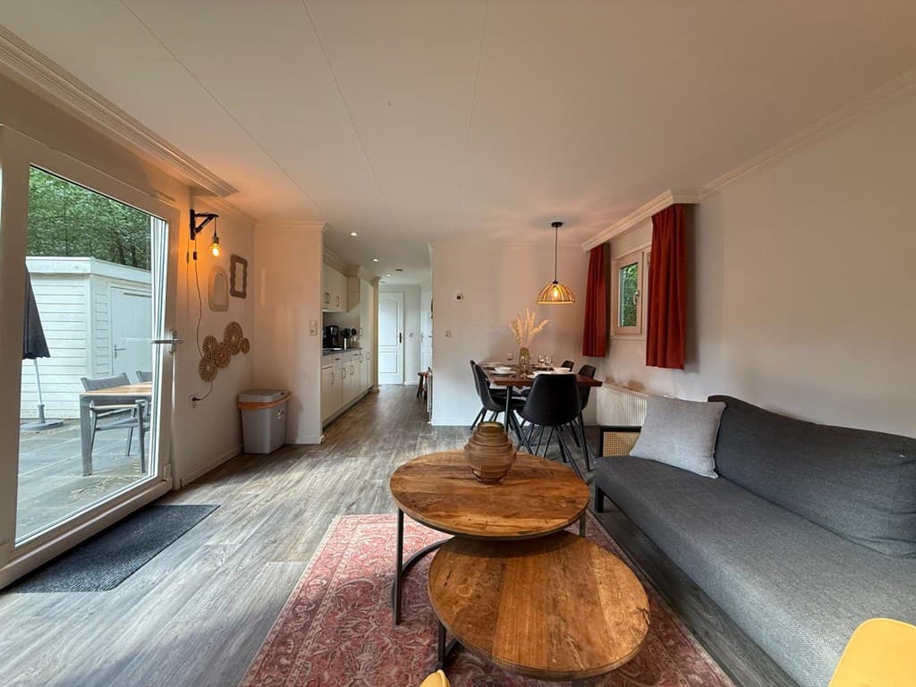 Zithoek met grijze bank, houten salontafels, eettafel en open keuken met lichte wandkasten en gordijnen.