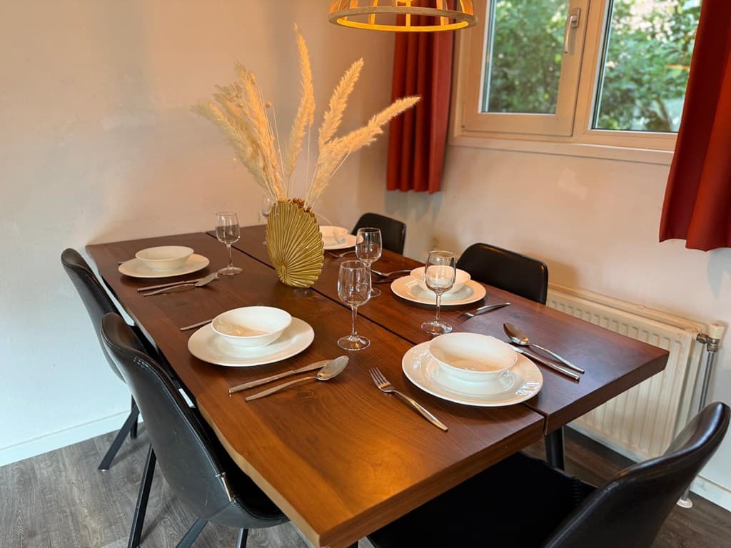 Eettafel met vier stoelen, gedekte tafel met borden, glazen en bestek, achter raam met uitzicht op groen.