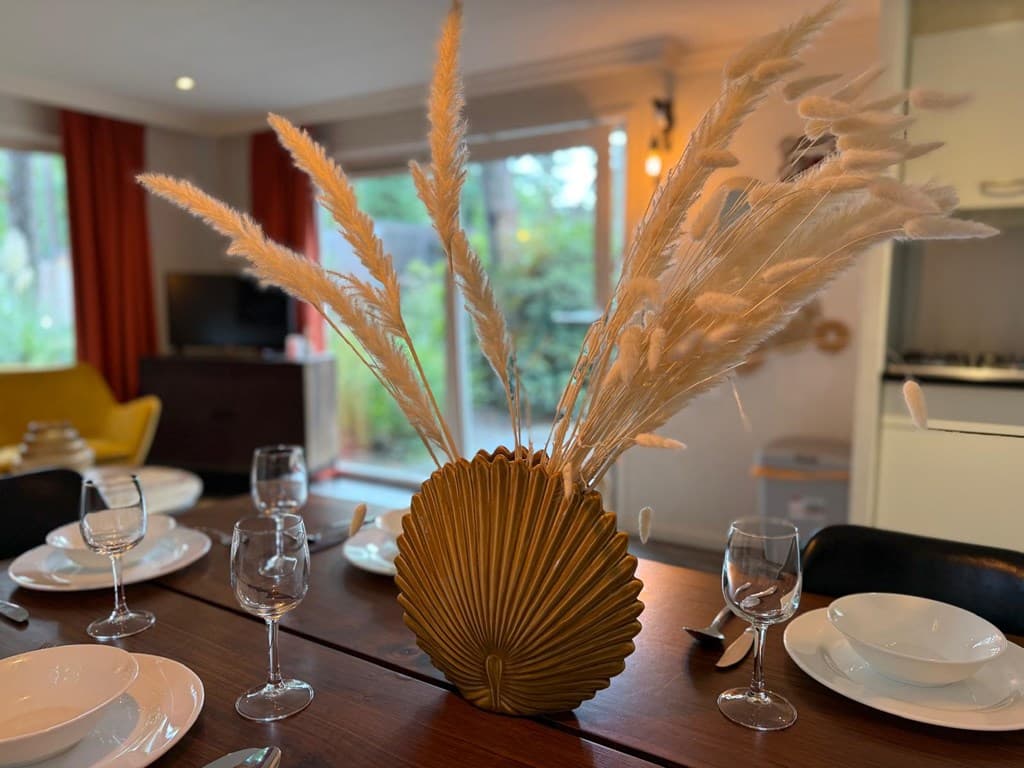 Eetkamer met een houten tafel, servies, glazen, en een gouden vaas met pluimen in een lichte ruimte met planten buiten.