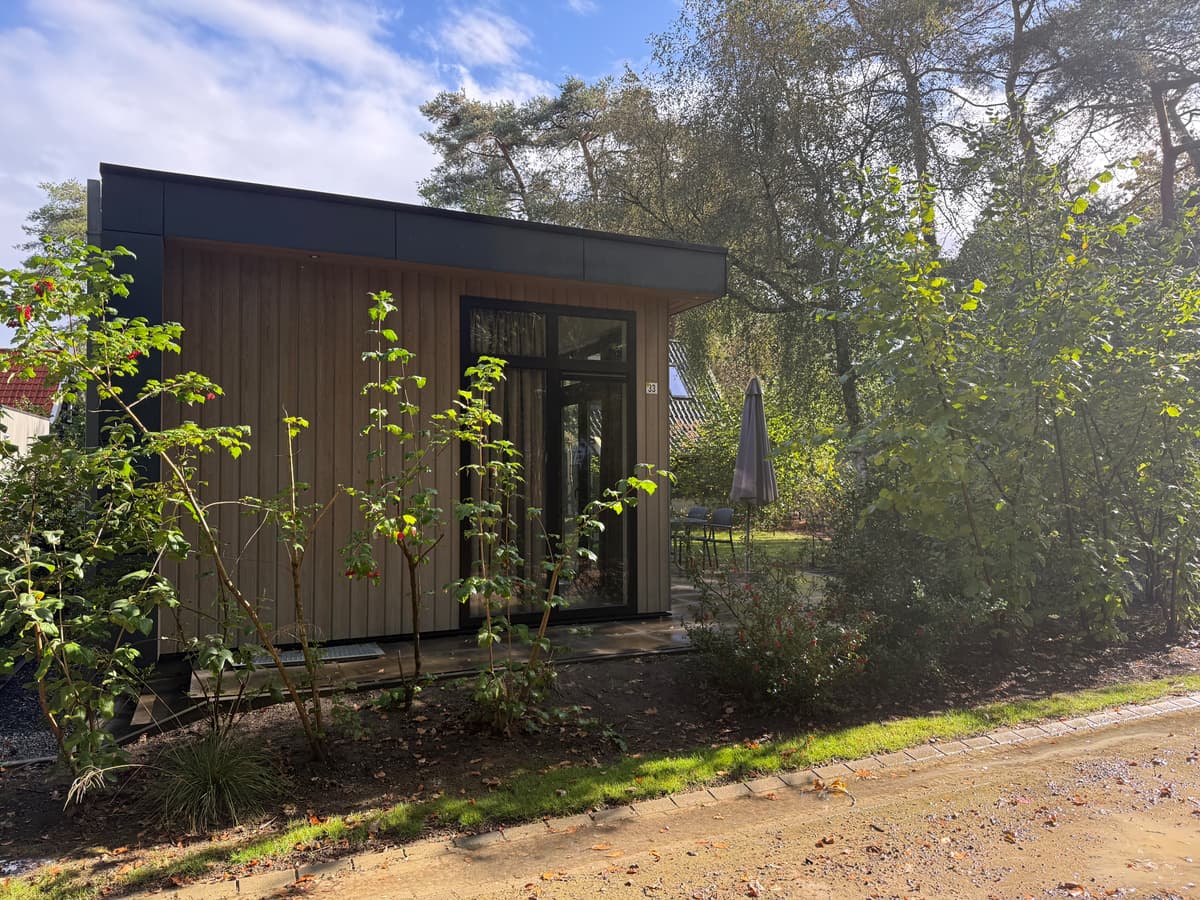 Moderne vakantiewoning met houten gevel, grote ramen en een terrasje naast een groene tuin in een bosachtige omgeving