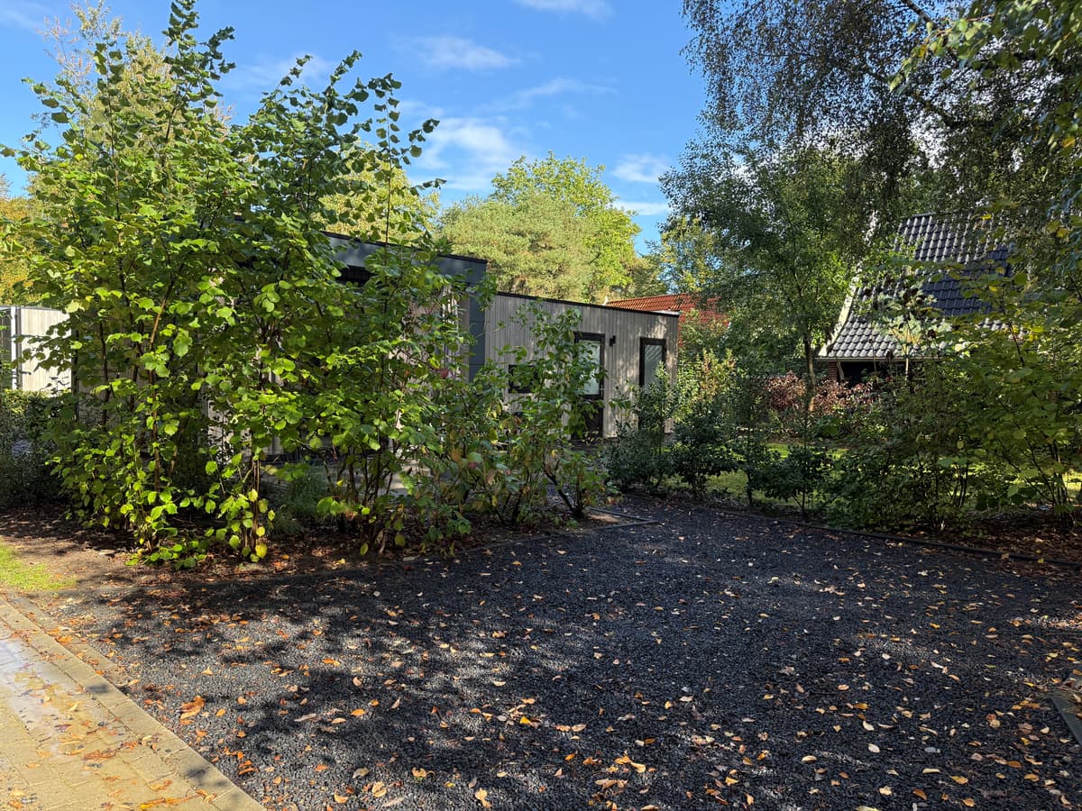 Moderne woning omgeven door groene planten en bomen met een grindpad en helder blauwe lucht erboven