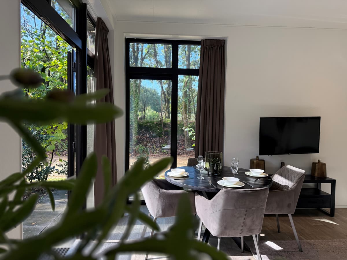 Eetgedeelte met een ronde tafel en stoelen, grote ramen met uitzicht op groen omlijst door bomen, televisie aan de muur