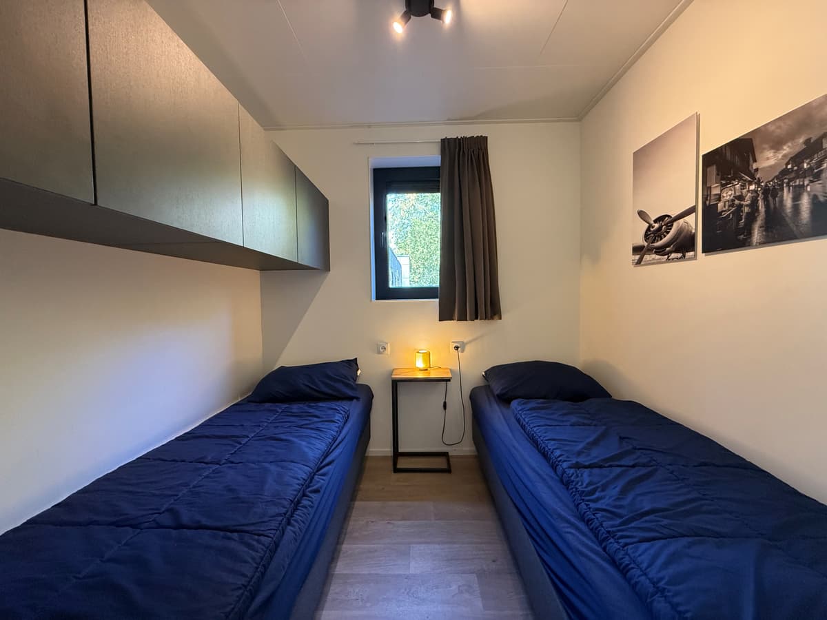 Twee eenpersoonsbedden met donkerblauwe beddengoed in een slaapkamer met muurkasten en een raam met uitzicht naar buiten