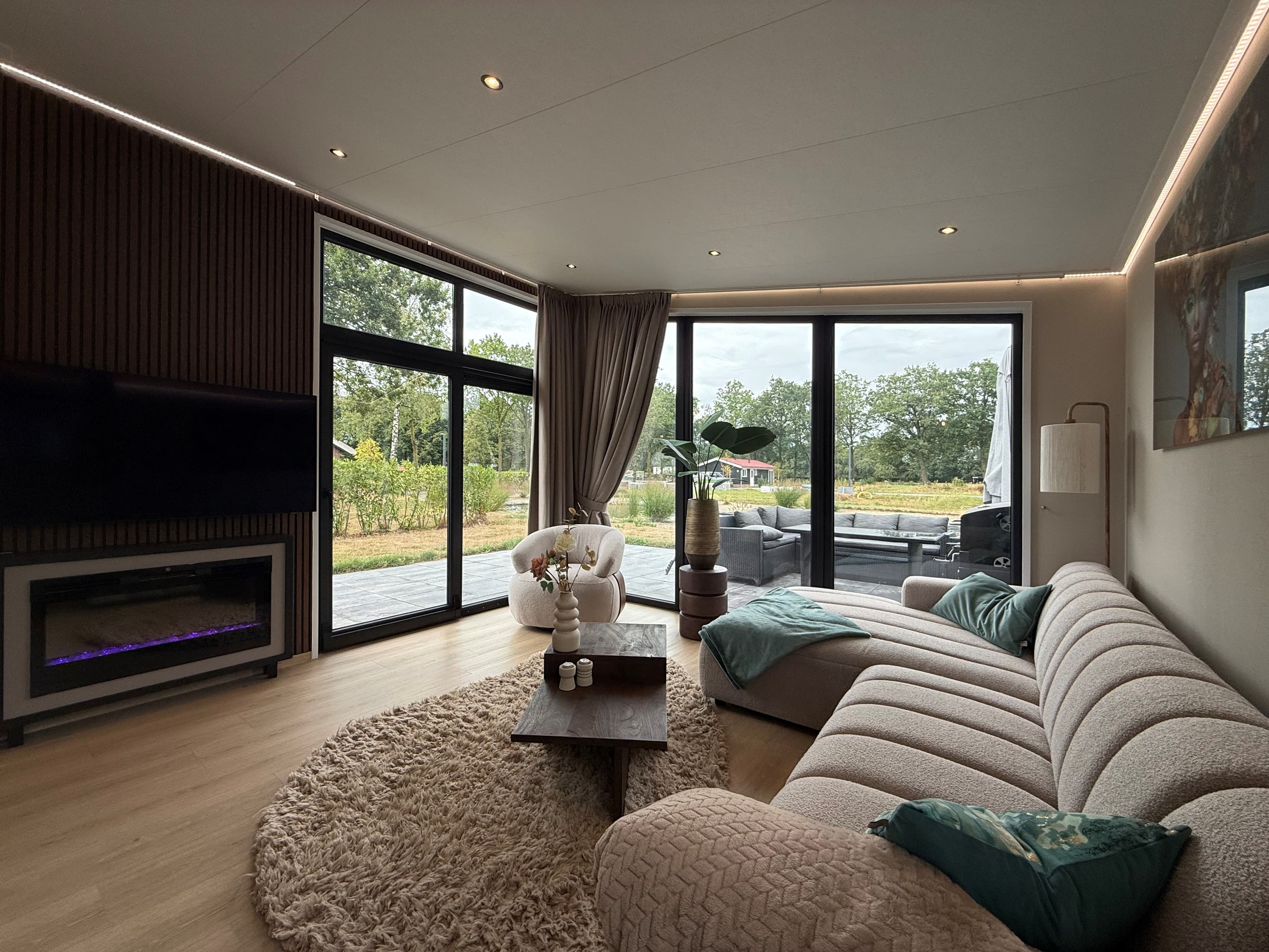 Lichte woonkamer met grote ramen, comfortabele bank, vloerkleed en zicht op het groene buitengebied.
