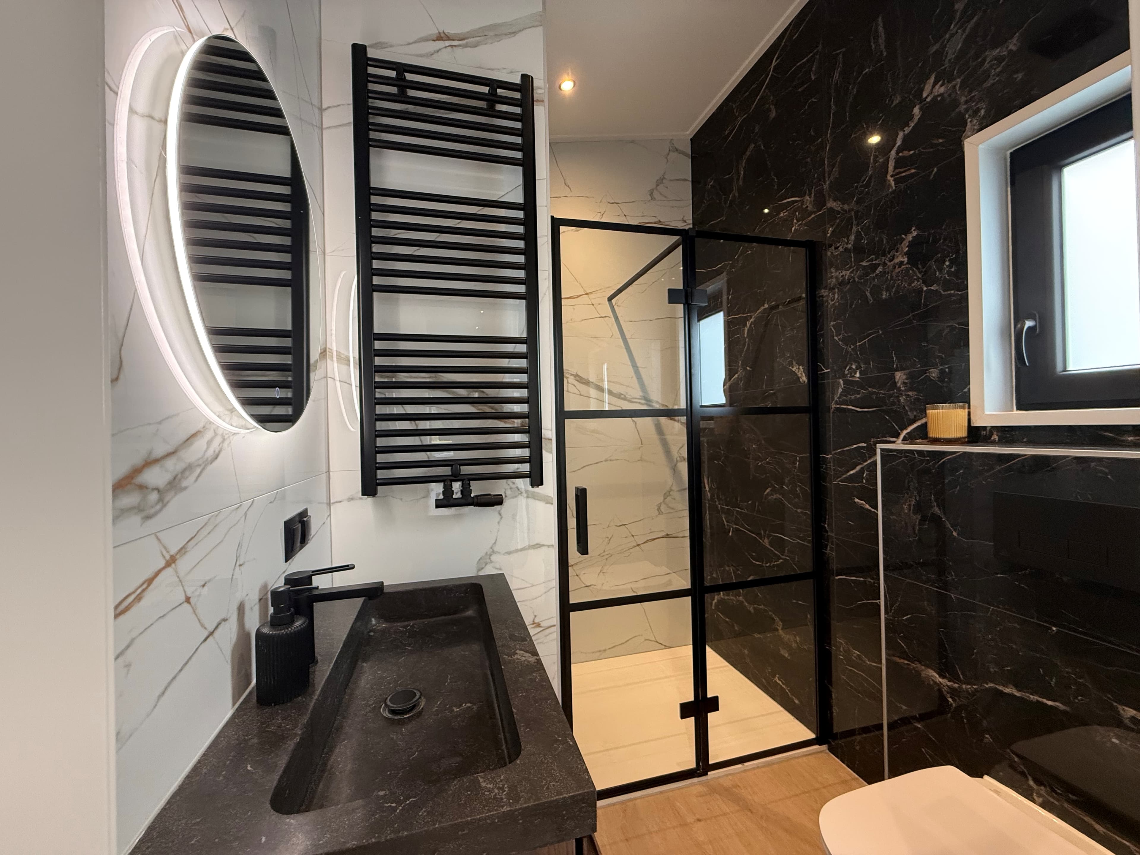 Moderne badkamer met zwarte marmerlook wand, inloopdouche met glazen wand en een ronde spiegel boven het wastafelmeubel.
