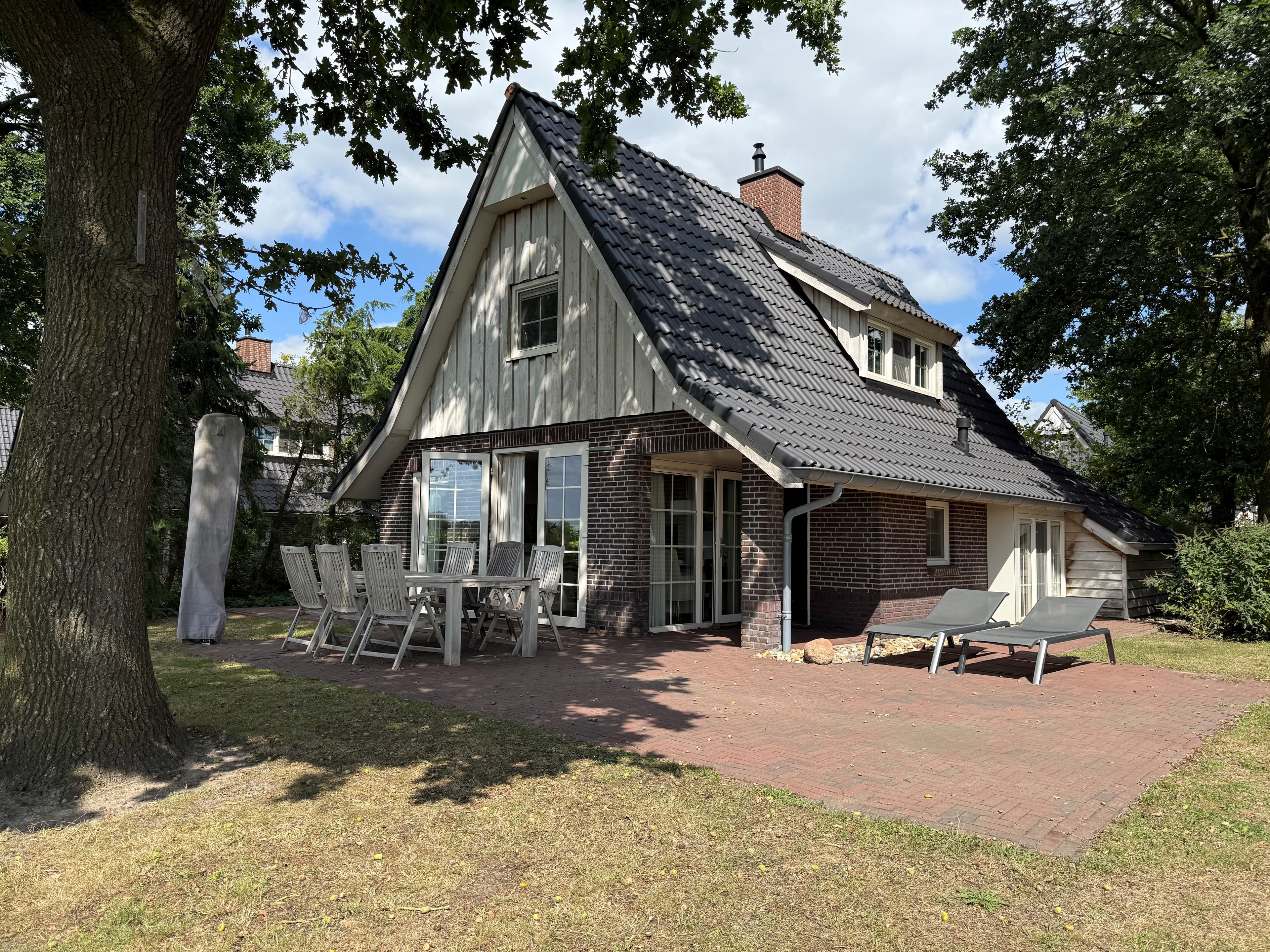 Recreatiewoning met glazen schuifpui, een terras met tafel en ligstoelen, omringd door bomen en gras.