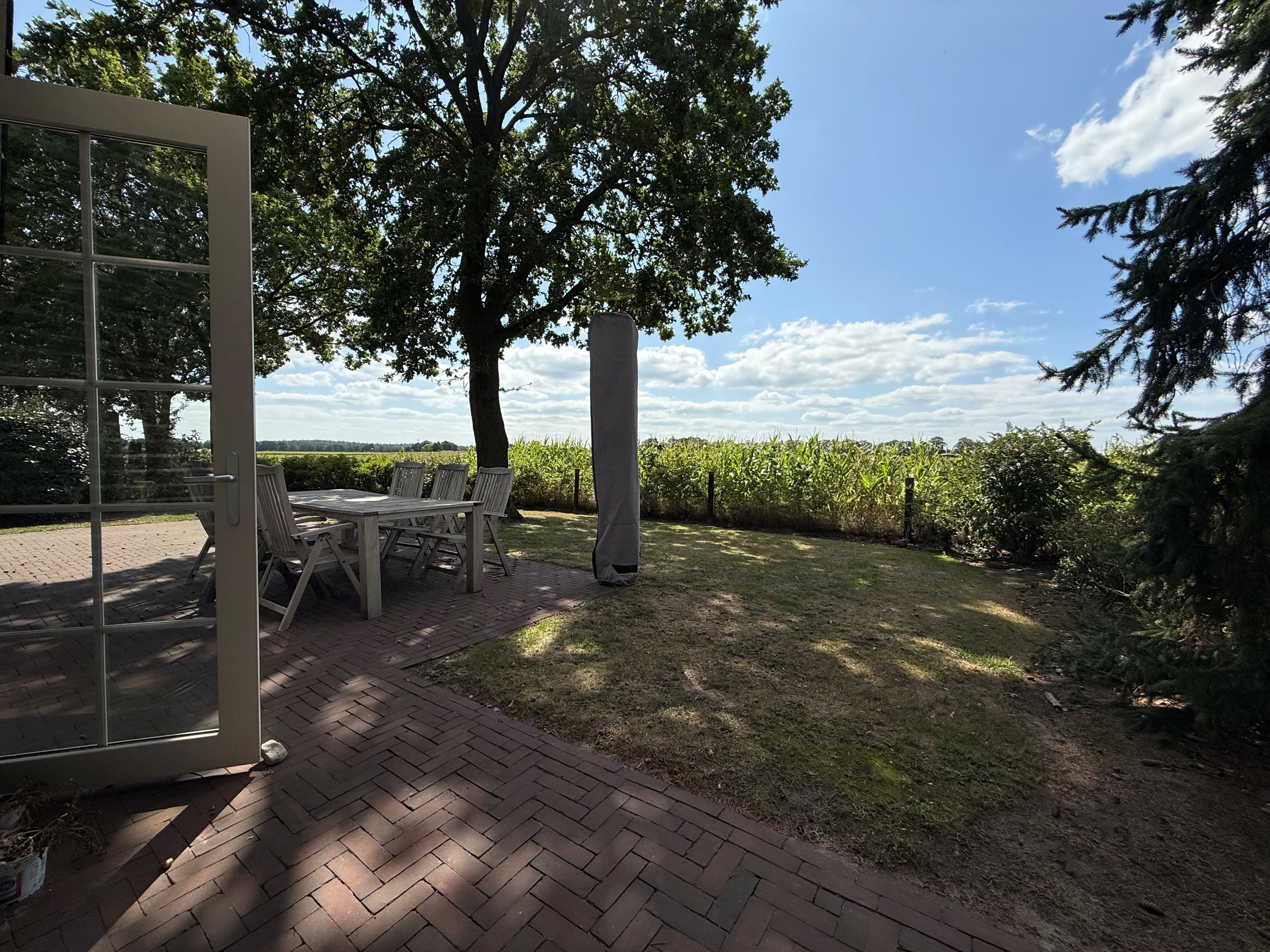 Tuin met uitzicht op groene akkers, houten terras met tuintafel en stoelen, gekaderd door bomen en struiken.