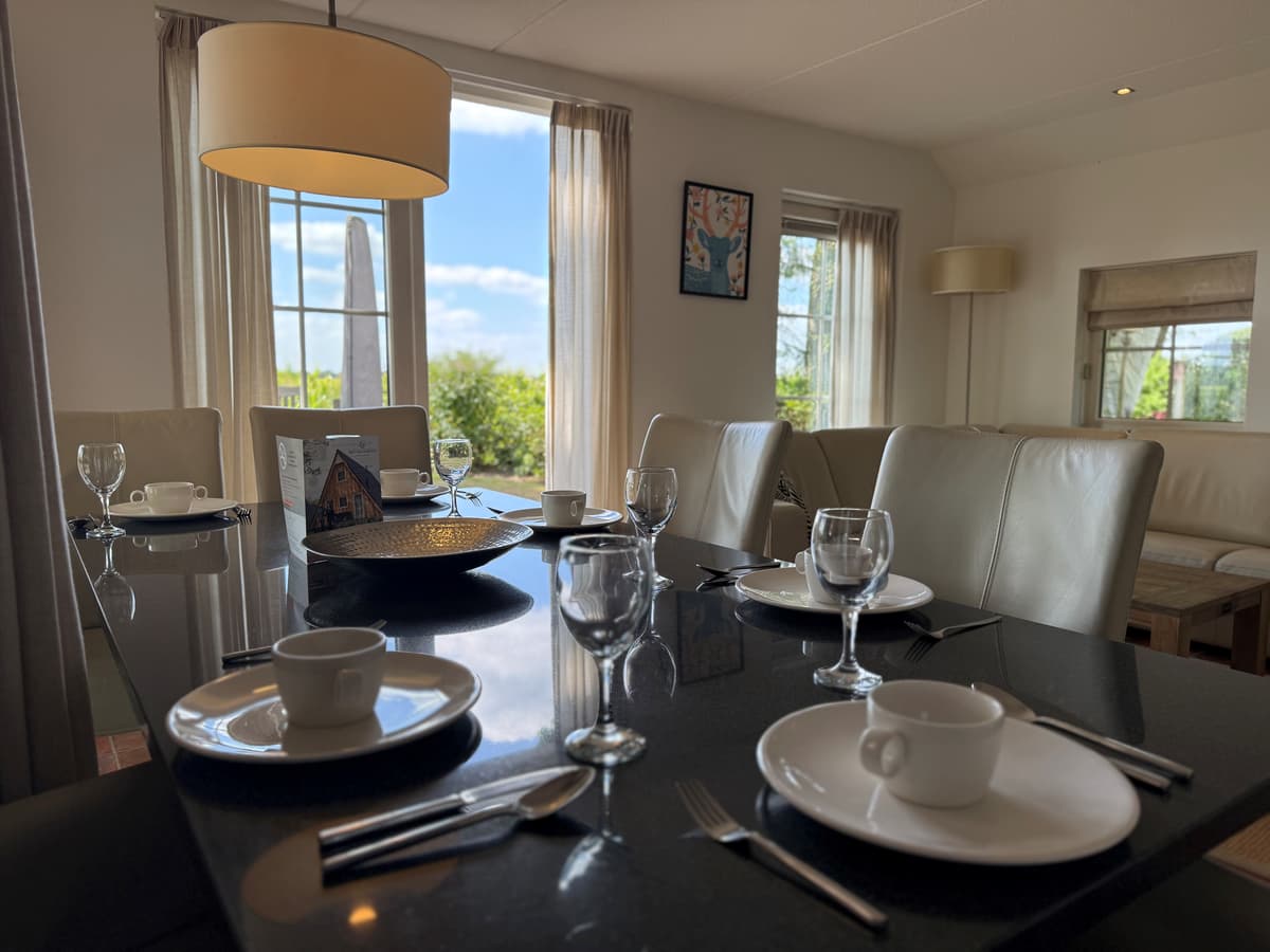 Eettafel met gedekte plaatsen, lichte meubels en grote ramen met uitzicht op groen vanaf de recreatiewoning